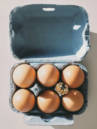 An open blue egg carton holding six brown eggs with a textured gold ring resting between them against a neutral background.