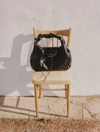 Black glossy leather handbag resting on a wooden chair against a white stucco wall in direct sunlight.
