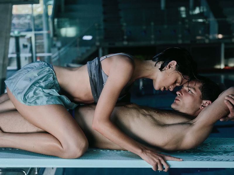 Adult couple lying on a diving board, embracing and looking at each other in a close-up shot with blue and grey hues.