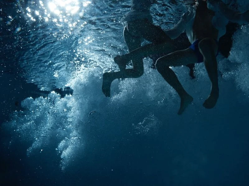 Underwater view of two children swimming, legs visible, in a blue swimming pool with the sun shining above the surface.