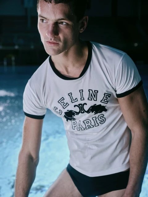 Athletic man in a white 'CELINE PARIS' t-shirt and black swim briefs poses near blue water, capturing a sporty, fashion-forward moment.