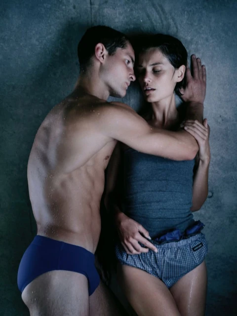 A couple, an adult man and woman, lie on a gray surface in swimwear, the man shirtless and the woman in a tank top and patterned shorts.