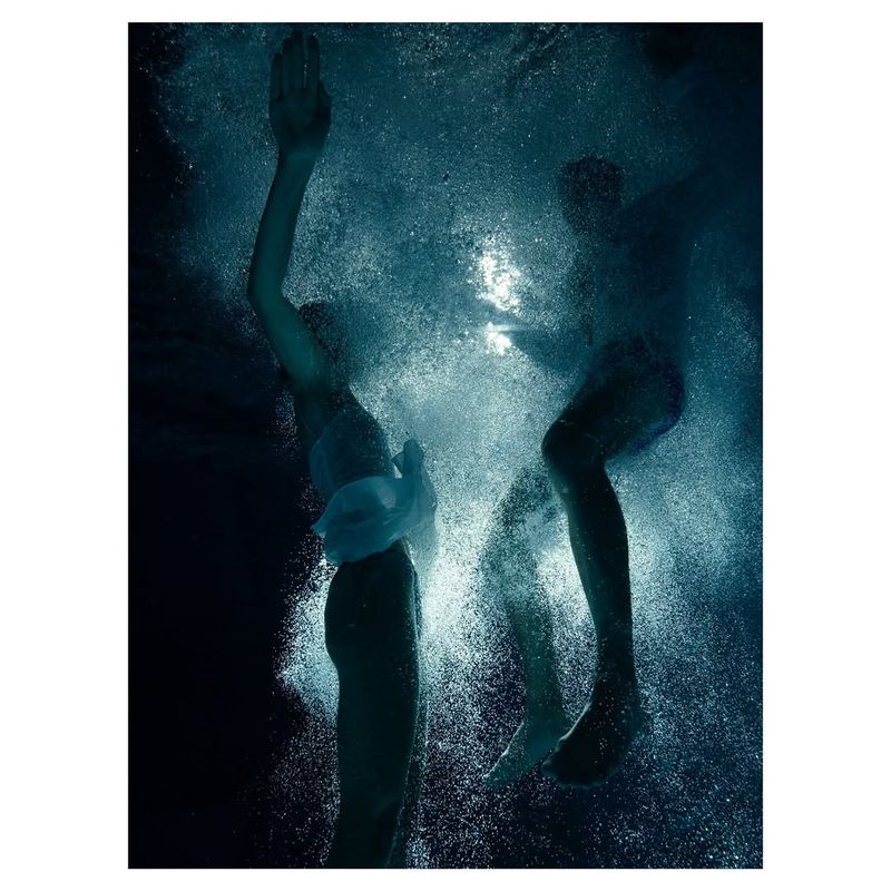Two people swim underwater, surrounded by bubbles in dark blue water, captured in a high-contrast, moody underwater photograph.
