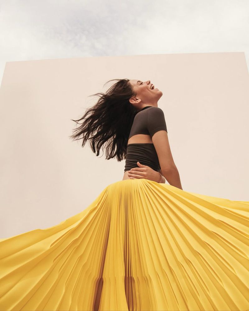 Smiling woman with dark hair and gray top with yellow pleated skirt posing against a wall and sky background in low-angle view.