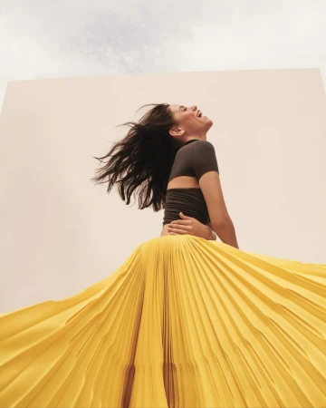Smiling woman with dark hair and gray top with yellow pleated skirt posing against a wall and sky background in low-angle view.