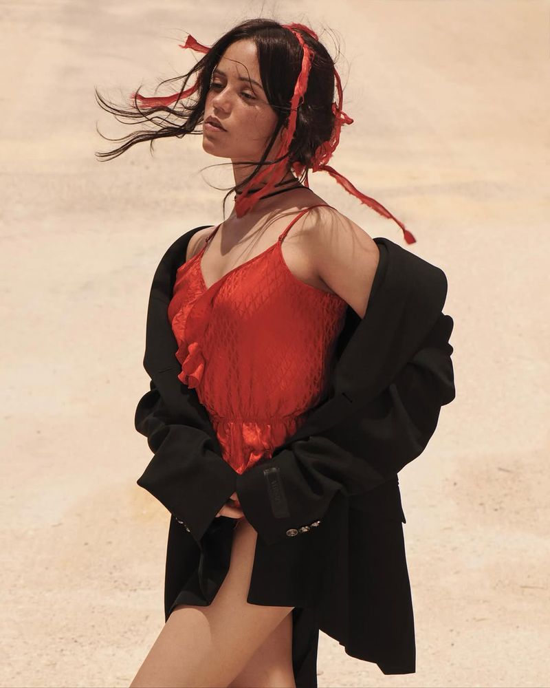 A woman in a red dress and black jacket poses in a sunlit outdoor setting with flowing red ribbons in her hair.