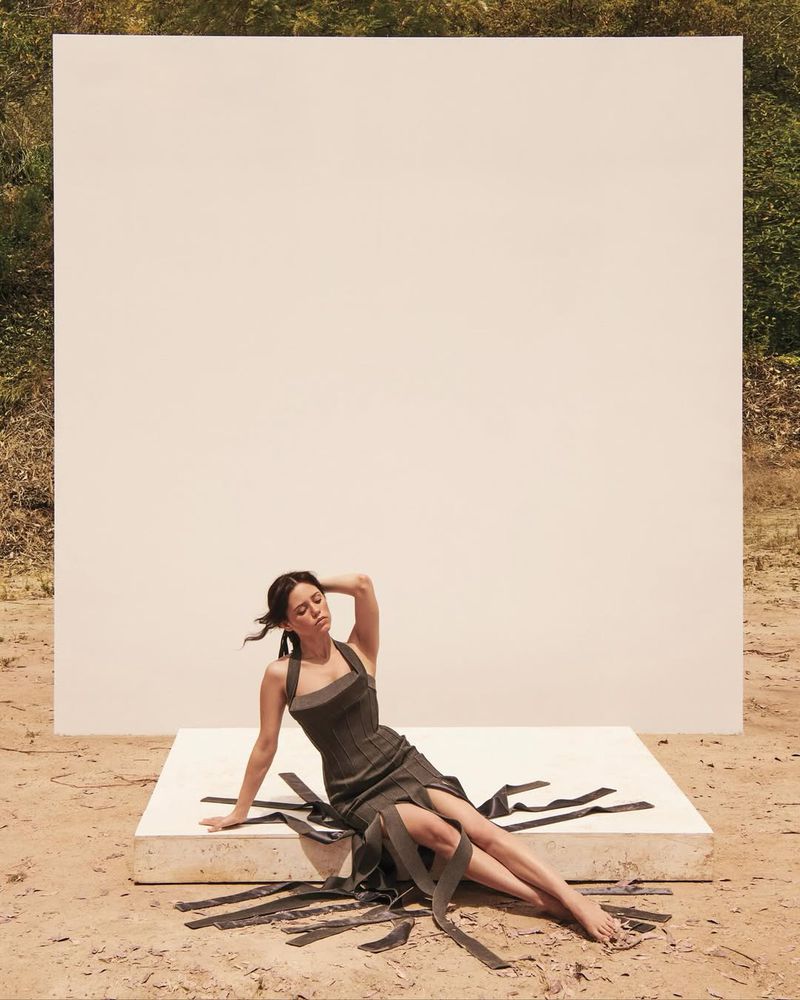 Young woman poses in a dress on a white surface in a field, with a large white square as a backdrop and tree line in background.