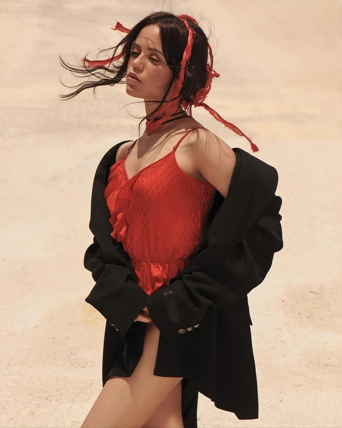 Jenna Ortega in a red top and oversized black jacket against a sandy outdoor background.