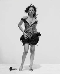 Jenna Ortega poses in an off-the-shoulder corset minidress with a ruffled skirt on a white studio floor with one shoe removed.
