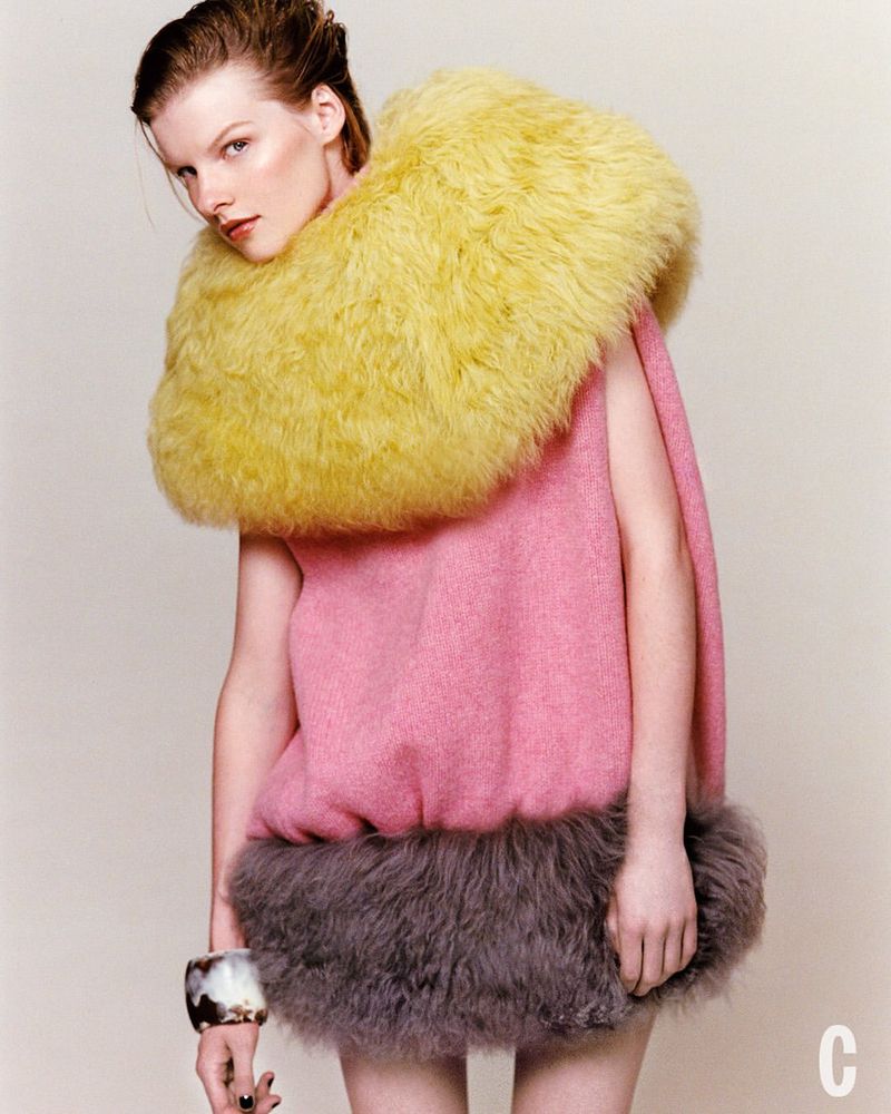A fair-skinned young woman poses in a pink sleeveless dress with yellow faux fur collar and gray faux fur hem, wearing a dark bracelet.
