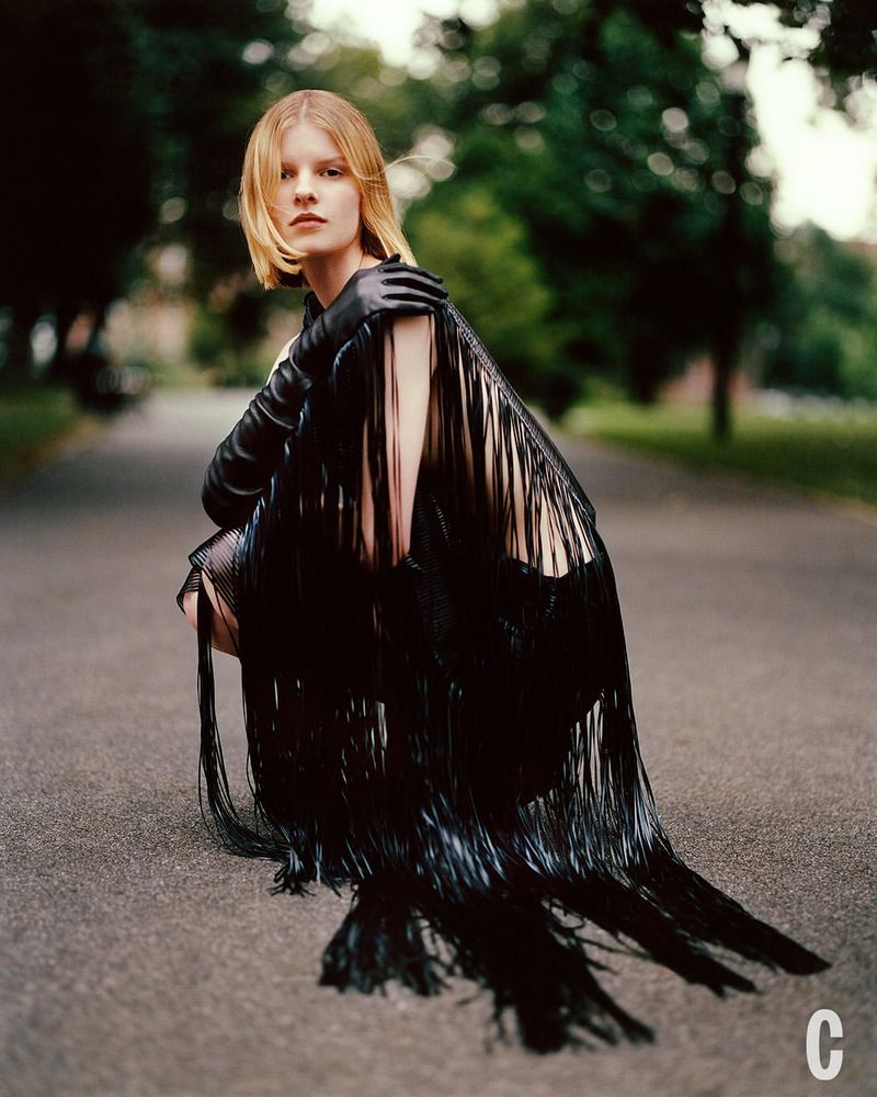 Fashionable woman in black fringe dress squats on a path outdoors, surrounded by greenery. Medium shot with natural lighting.