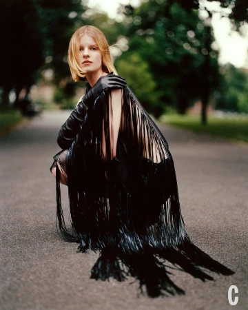 Fashionable woman in black fringe dress squats on a path outdoors, surrounded by greenery. Medium shot with natural lighting.