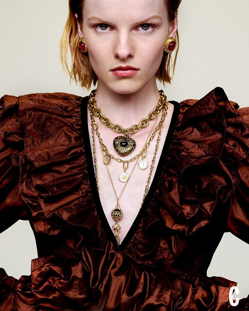 A woman in a brown ruffled dress and gold jewelry poses in a fashion editorial shot with a plain backdrop and a medium shot.
