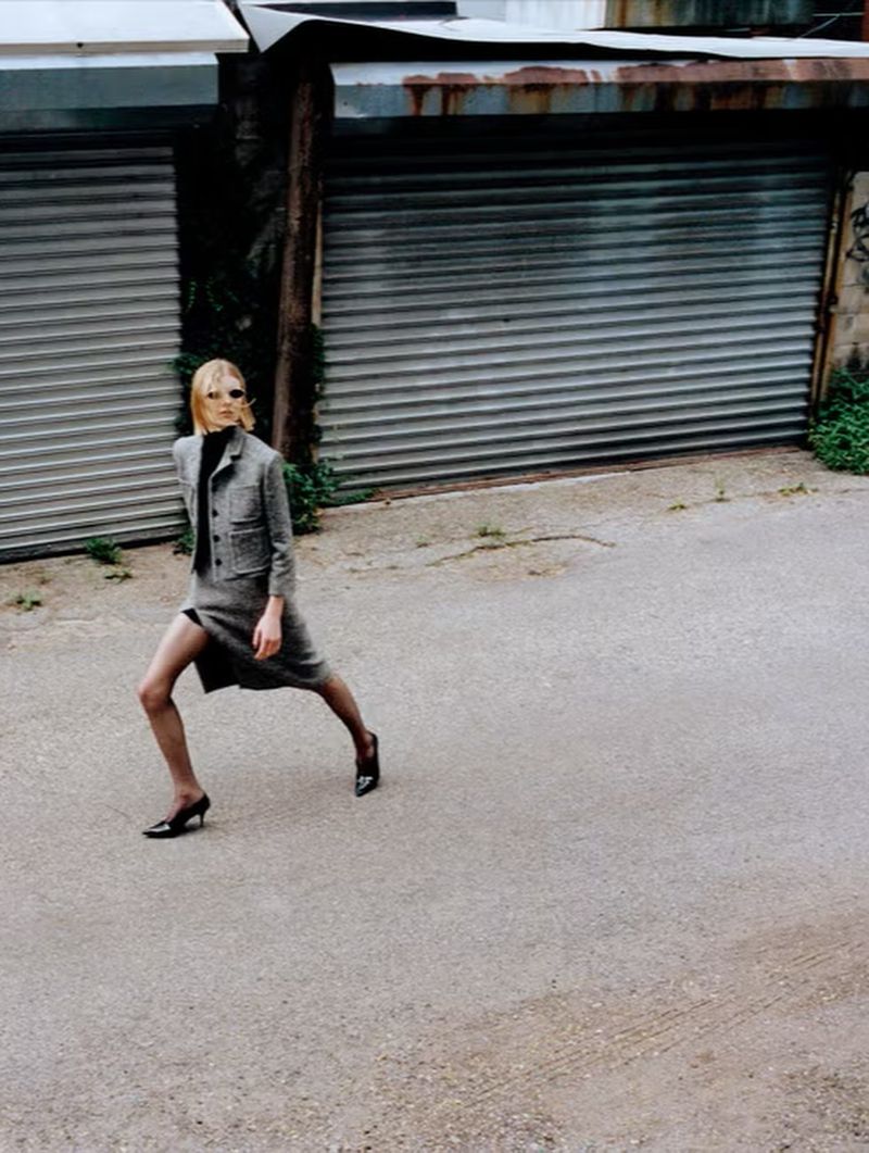 A woman in a grey skirt suit and black heels confidently walks down an alleyway with weathered rolling doors on either side, fashion shoot.