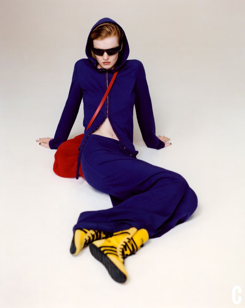 Fashion shot of a person in blue hooded outfit and black sunglasses sitting on white background with red bag and yellow/black boots.