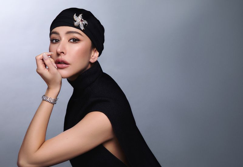 Elegant portrait of a woman wearing a black headscarf with a jeweled pin and a matching turtleneck sweater against a gradient grey backdrop.