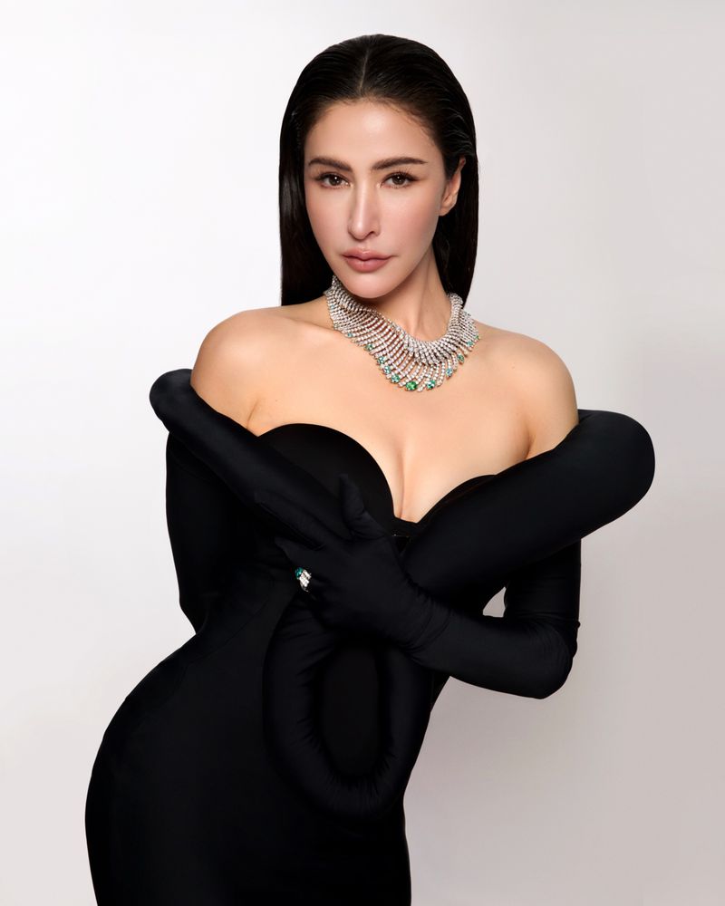Sririta Jensen wearing a black off-the-shoulder gown with attached gloves and an elaborate diamond necklace against a studio background.