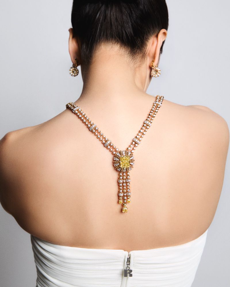 Woman showcasing a gold necklace and earring jewelry set with yellow gemstone accents, wearing a white strapless dress against a grey background.