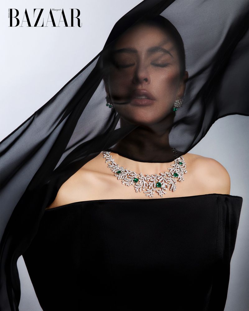 Woman in black dress with diamond and emerald necklace, partially veiled with sheer fabric, posing in front of light gray background.