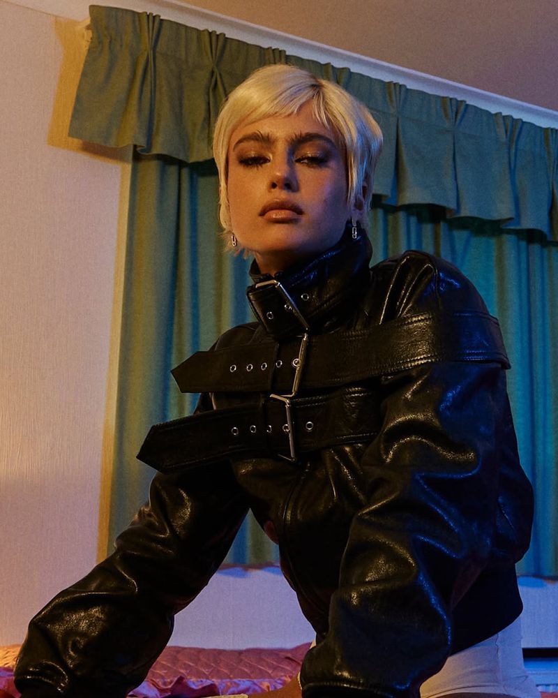 Stylish woman with short blonde hair poses in black leather jacket with silver buckles in an indoor setting with vintage curtains.