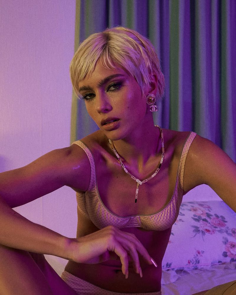 Portrait of a woman with short blonde hair in lingerie sitting on a bed with floral sheets in an indoor environment with pink lighting.
