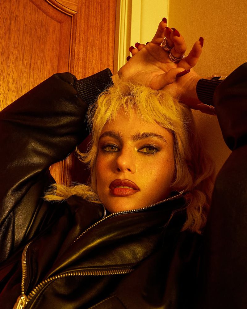 Portrait of a young woman with blonde hair wearing a black leather jacket, posing indoors with her hands above her head.