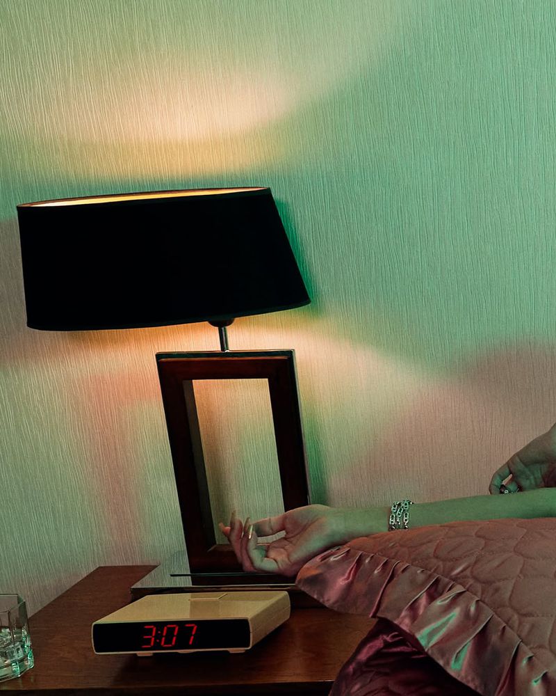 Still life photo of a retro digital alarm clock on a wooden nightstand next to a lamp, a woman's arm, and a glass, bathed in green and orange light.