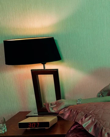 Still life photo of a retro digital alarm clock on a wooden nightstand next to a lamp, a woman's arm, and a glass, bathed in green and orange light.