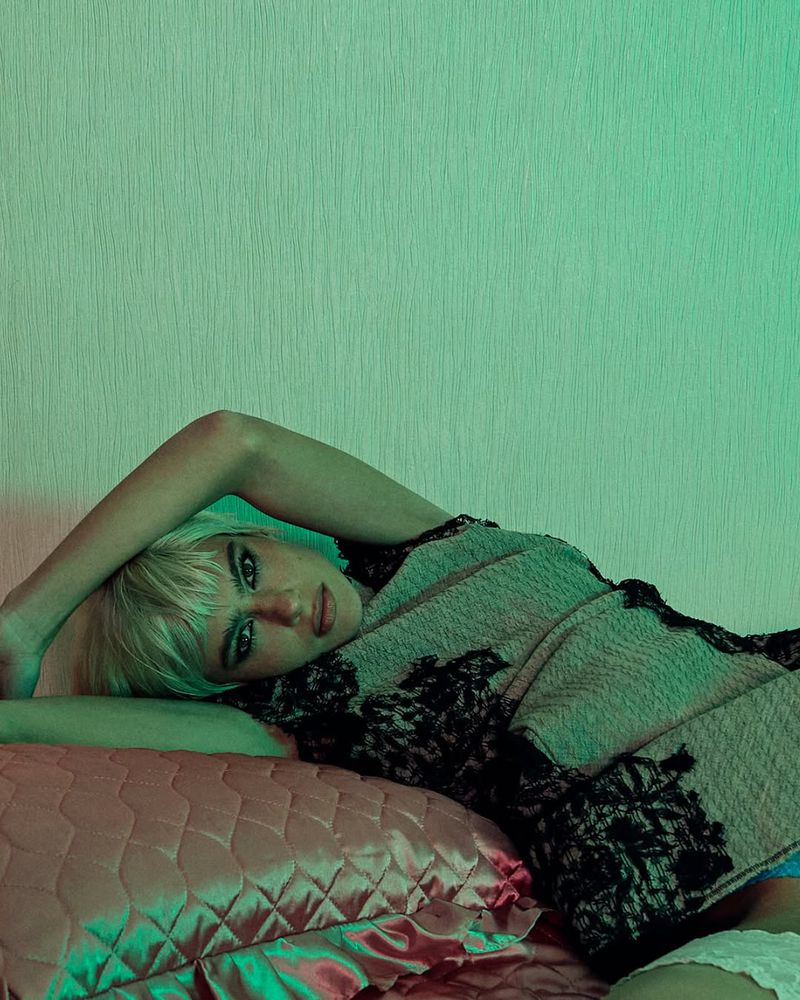 Blonde woman lies on a pink quilted pillow and bed in a dimly lit indoor setting with a textured wall in the background.