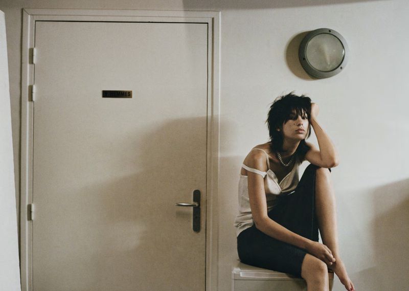 A young adult woman with dark hair, wearing a white tank top and black pants, sits in a light-filled indoor setting with a door in the background.