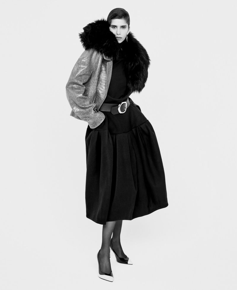 Elegant studio portrait of a woman wearing a black dress and a metallic jacket with fur collar against a white backdrop.