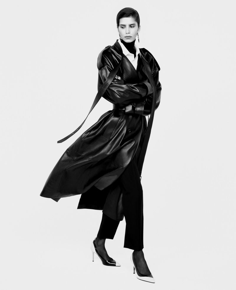 Black-and-white fashion portrait of a woman in a stylish black leather trench coat, black pants, and two-tone heels against a white background.