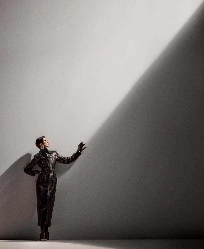 Elegant woman in a black leather outfit poses against a gradient gray background with dramatic lighting and shadows in a studio setting.