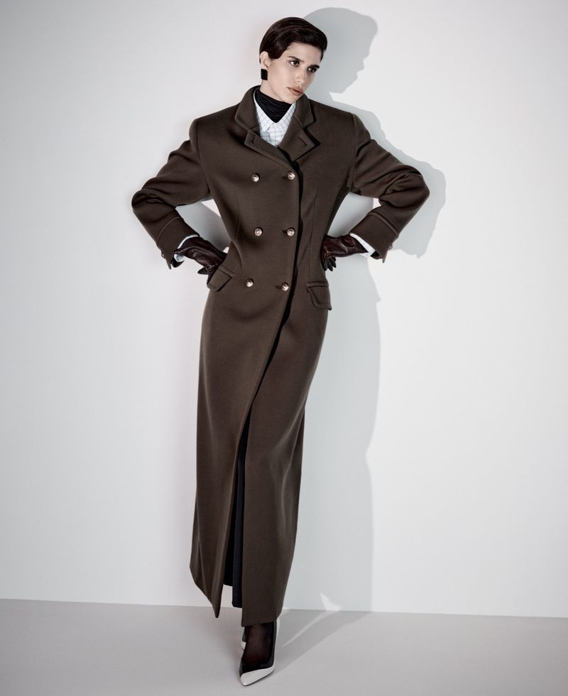 A model poses in a long brown double-breasted coat with black gloves, tights, and heels against a plain white backdrop in a studio setting.