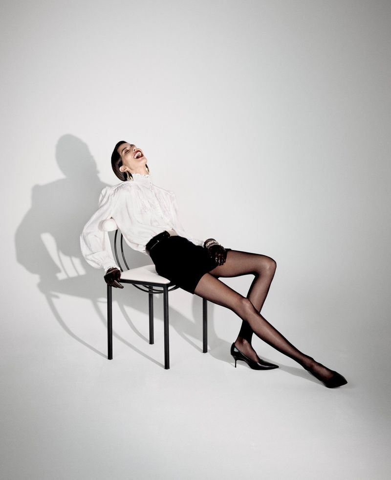 A woman in a white top, black skirt, and black heels sits on a chair against a white backdrop, head tilted back and laughing.