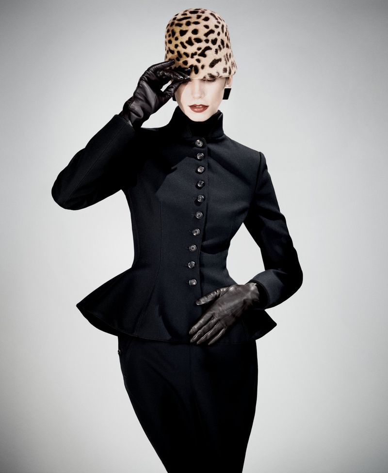 Fashionable adult female poses in a black peplum suit with gloves and a leopard print hat against a pale gradient backdrop in studio lighting.