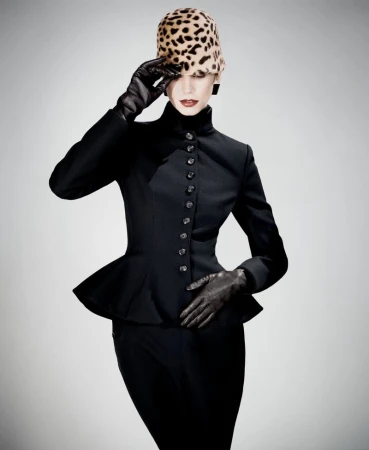 Fashionable adult female poses in a black peplum suit with gloves and a leopard print hat against a pale gradient backdrop in studio lighting.