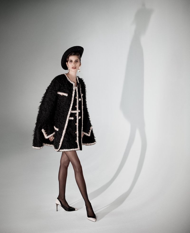 Model wearing a textured black tweed cape coat with white trim, black hat, tights, and two-tone slingback heels against a white backdrop.