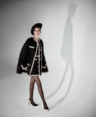 Model wearing a textured black tweed cape coat with white trim, black hat, tights, and two-tone slingback heels against a white backdrop.