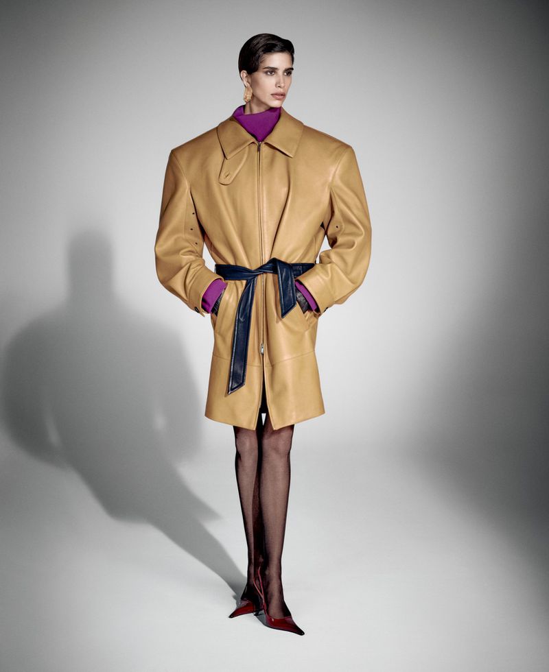 A fashionable woman posing in an oversized tan leather trench coat, worn with a navy belt and purple turtleneck against a gradient grey backdrop.