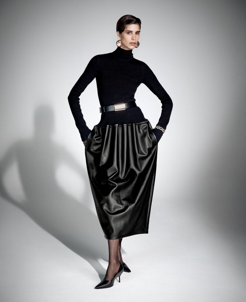 Elegant woman poses in a black turtleneck, belted black skirt, gold hoop earrings, and black heels against a gradient white background.