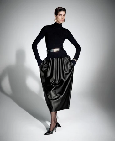 Elegant woman poses in a black turtleneck, belted black skirt, gold hoop earrings, and black heels against a gradient white background.