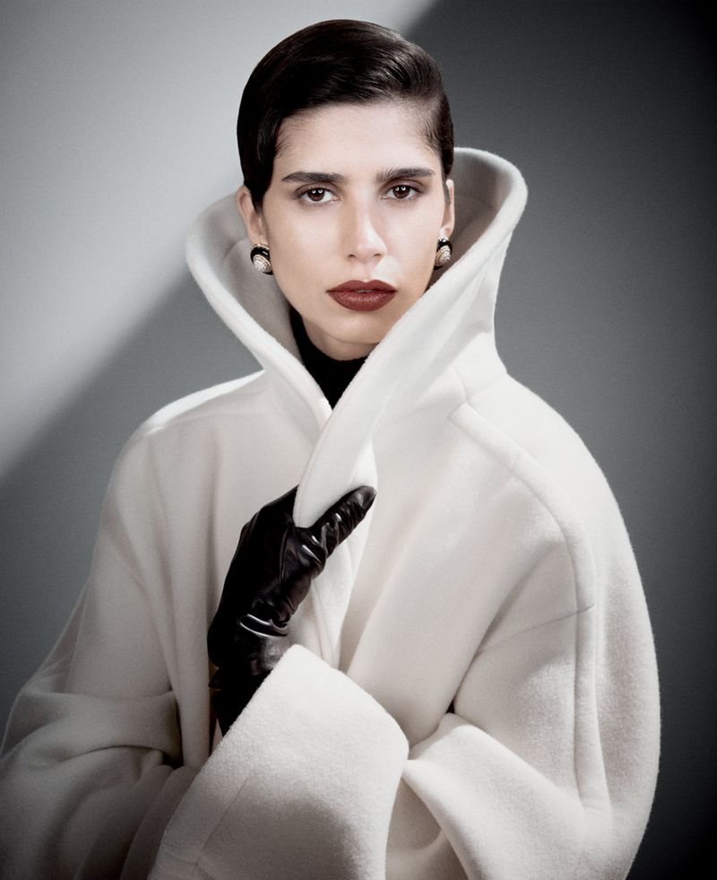 Fashionable woman with dark hair, dark lipstick, and pearl earrings poses wearing an elegant white coat against a grey background.