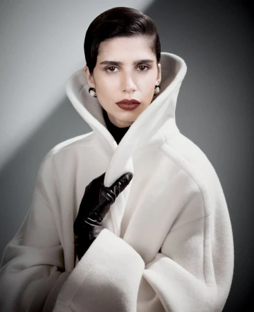Fashionable woman with dark hair, dark lipstick, and pearl earrings poses wearing an elegant white coat against a grey background.