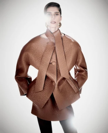 Model Mica Argañaraz wearing a structured brown textured leather jacket with an attached neck scarf against a glowing backdrop.