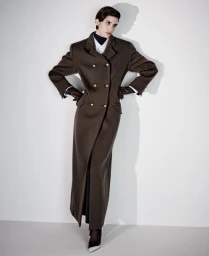 Model Mica Argañaraz posing in a long brown double-breasted maxi coat and gloves against a white studio wall with a cast shadow.