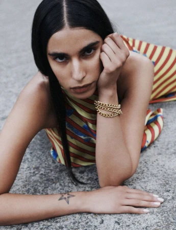 Woman lying down wearing a yellow and red striped dress with a gold chain bracelet, resting her head on her hand outdoors.