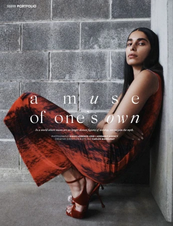 Adult female model poses leaning against a grey block wall in an indoor fashion shoot. She wears a red printed dress and high heels.