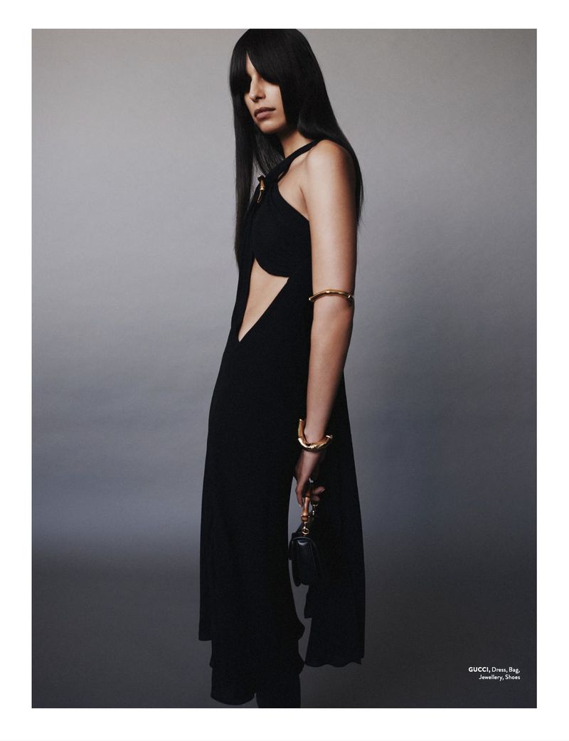 Elegant woman in a black dress with cut-out stands in a photo studio, showcasing gold jewelry and a small black purse.