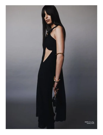 Elegant woman in a black dress with cut-out stands in a photo studio, showcasing gold jewelry and a small black purse.